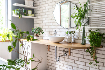 Fototapeta na wymiar Interior of bathroom in industrial style with white bricks on the wall, stylish mirror, green plants, wooden counter with ceramic sink. Spa with plant and window.
