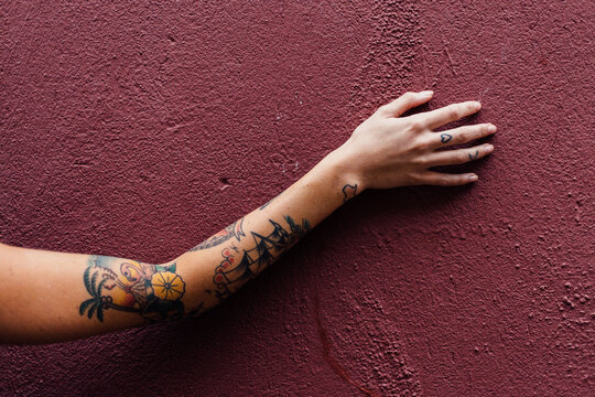 Tattooed arm reaching up a maroon concrete wall.