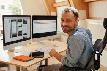 Portrait of programmer smiling at camera while sitting at his workplace with computer monitors and...