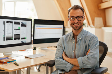 Portrait of successful software developer in eyeglasses standing with arms crossed and smiling at...