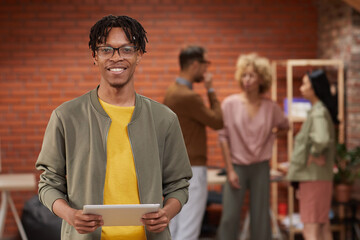 Portrait of young African designer holding tablet pc and smiling at camera with his colleagues standing in the background