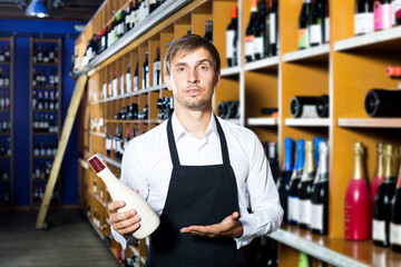 Positive young man seller wearing uniform having bottle of wine in wine house