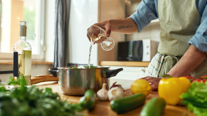 Cropped shot of man, Italian cook pouring a glass of white wine into the pan with chopped vegetables while preparing a meal in the kitchen