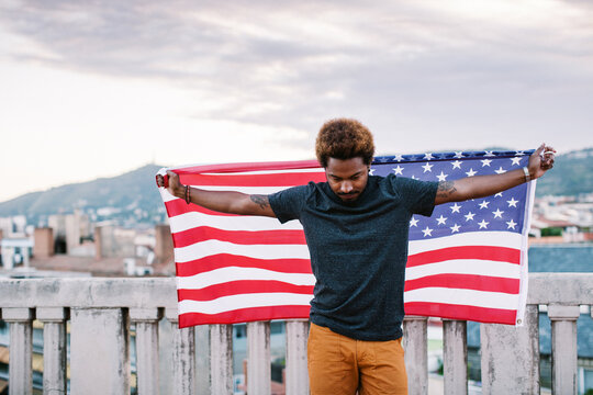 Young black man holding an american flag standing on a rooftop.