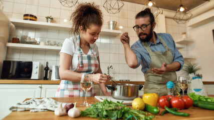 Italian man adding pepper, spice to the soup while woman stirring it with a spoon. Couple preparing a meal together in the kitchen. Cooking at home, Italian cuisine