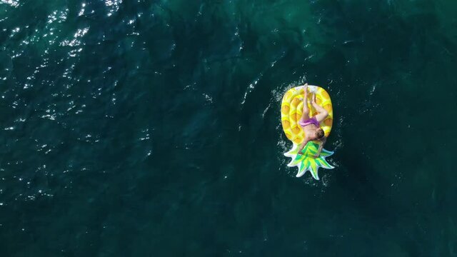 Aerial view of a guy in pink shorts who swims in the sea on a pineapple-shaped inflatable mattress. The camera moves up.
