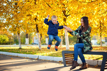 Obraz premium Beautiful mother and son play on a bench in the autumn Park. The boy jumps off the bench. Relationships between parents and children. Mother's day
