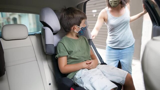 Caucasian child sitting in the car seat with his protective mask on. His mother opens the door and fastens his seat belt. Concept new rules, new normality, coronavirus pandemic.
