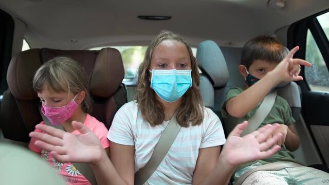 Caucasian Teenager And Two Kids In The Back Seats Of A Car With Protective Masks On. They Are Dancing. Concept New Rules, New Normality, Coronavirus Pandemic.