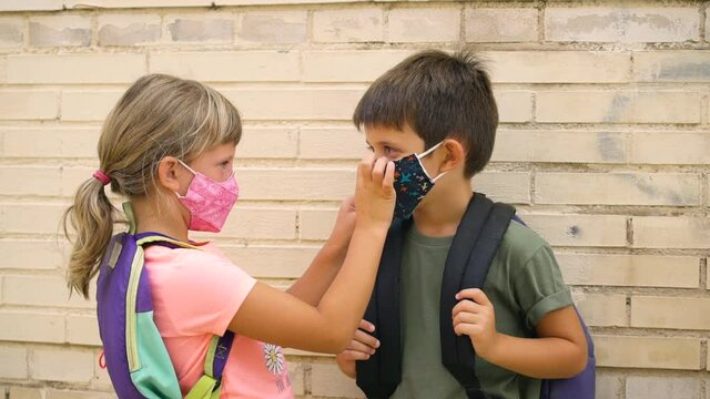 Elementary students with backpacks and protective face masks. Caucasian girl helps a boy to put his face mask correctly outside school. First day of school during Coronavirus pandemic.