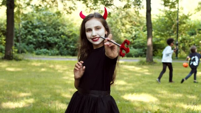 Medium shot of joyful Caucasian girl wearing devil costume is standing outdoors, looking at camera and smiling while other kids playing on background
