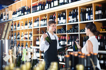 Seller wearing apron helping to buy bottle of wine to woman customer in wine store