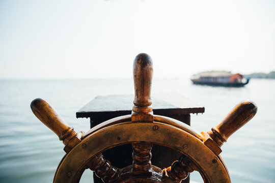 Old boat steering wheel in the sea with a house boat in the background. Kerala backwaters, India.