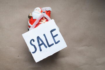 a small Christmas toy house in the snow with Santa Claus on the roof lies on a craft brown background with a sign discount Christmas toy on a brown background