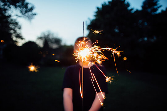a boy holding a sparkler in front of his face in the early evening