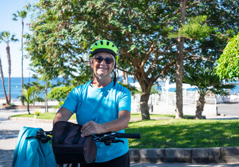 Senior active woman with electrice bicycle resting after excursion - public park with palm trees and horizon over water - healthy lifestyle for retired people