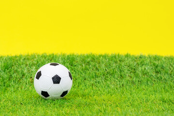 Fototapeta premium Miniature toy soccer ball lies on green grass of artificial turf of football field, yellow background, front view, close up. Equipment for outdoor activities and sports.