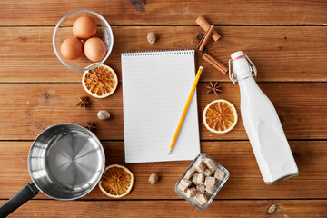 cooking, christmas and seasonal drinks concept - notebook with pencil, ingredients for eggnog, pot on wooden background