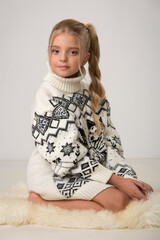 Beautiful little girl child in a big white sweater sits on a cube in the studio