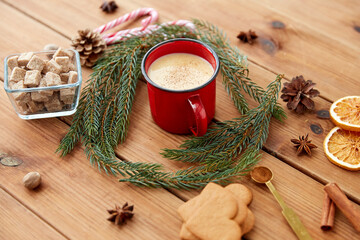 christmas and seasonal drinks concept - red cup of eggnog with cinnamon, fir tree branches, gingerbread, sugar and candy canes on wooden background