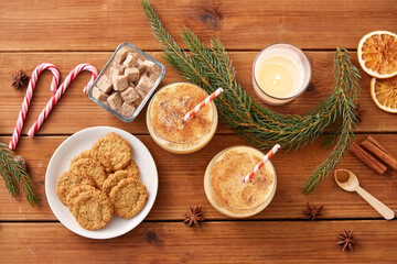 Obraz na płótnie Canvas christmas and seasonal drinks concept - glasses of eggnog with oatmeal cookies, candy canes, sugar, fir tree branches and candle burning on wooden background