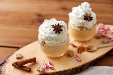 christmas and seasonal drinks concept - glasses of eggnog with whipped cream topping, candy canes, sugar and cinnamon on wood cut board