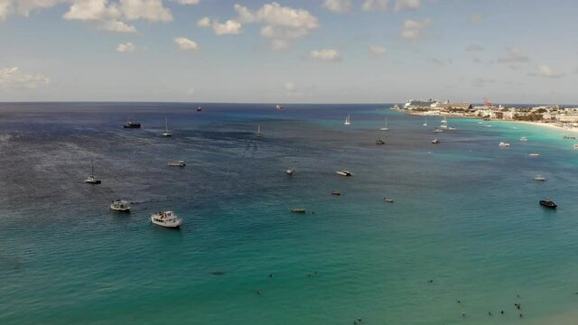 A beautiful aerial of the beach in Barbados. Drone is flying backward from the open blue sea with the boats over the beach and the palm trees.