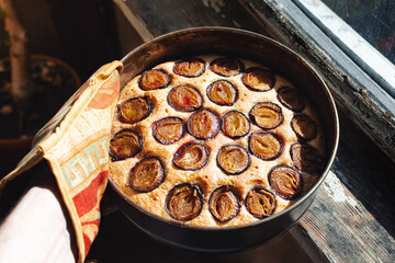 a hand holds a freshly baked plum pie in a baking dish by an old wooden window