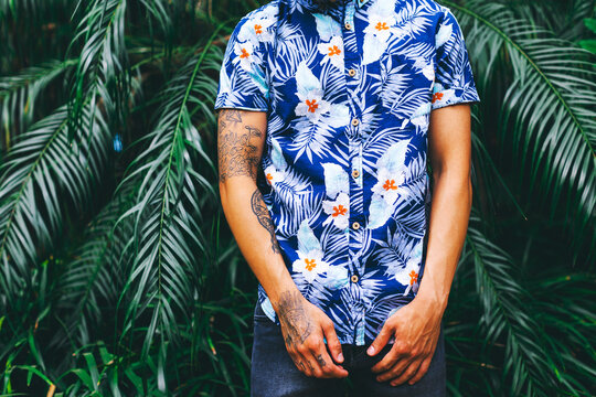 Closeup of a tattooed man wearing a flowery shirt standing in front of palm tree leaves.