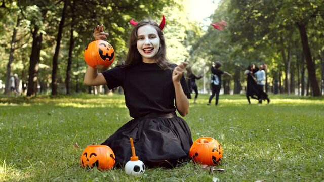 Wide shot of cheerful Caucasian girl wearing devil costume is sitting on grass outdoors, looking at camera and smiling while other kids playing on background