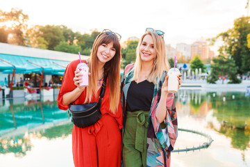Lifestyle portrait of two graceful smiling women in bright summer outfit spending holidays together, enjoying summer, walking  outdoor. Drinking sweet cocktails.