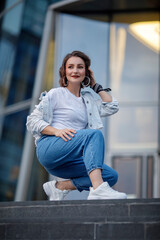 Fototapeta premium A beautiful girl in blue jeans and a white t-shirt is sitting on the stairs. Portrait of a pretty girl in a white jacket.
