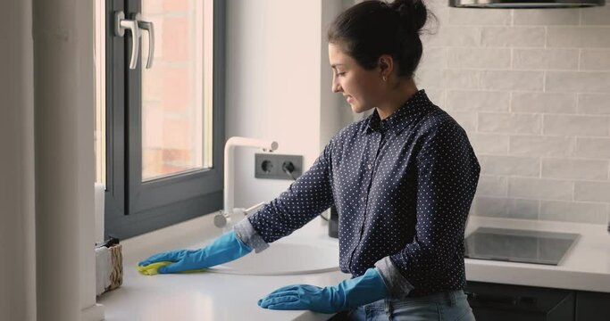 Young indian housewife home do chores wear rubber protective gloves use rag clean countertop surface of kitchen table enjoy process. House cleaning services worker accomplish work smile look at camera