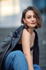 A beautiful woman in blue jeans, black top and jacket is sitting on the stairs. Portrait of a girl in a leather jacket. City portrait.