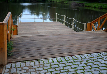 Fototapeta premium wooden fence made of beams painted brown and screwed into the shape of an x cross at the shore of a pond with a floating plank pier with a rope railing in a city park