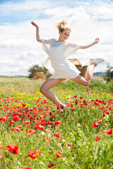 Pretty young female in white dress jumping in poppy field of wild flowers