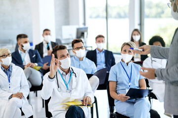 Happy doctor with protective face mask raising hand to ask a question while attending educational...