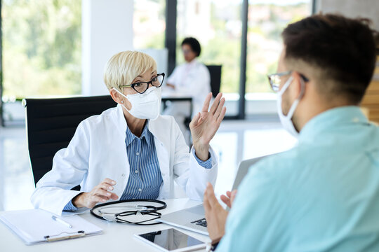 Mature Female Doctor With Face Mask Talking To A Patient At Medical Clinic.