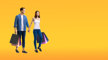 Holiday sales action, shop, consumer concept - couple with shopping bags, going for purchases, holding hands. Isolated over orange yellow background. Full body length studio portrait. Copy space area.