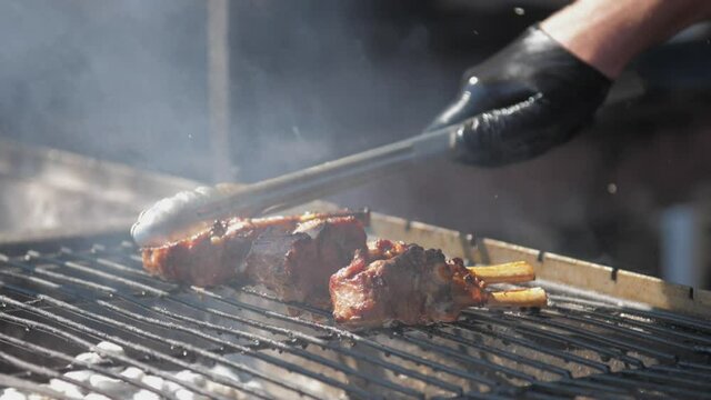 Person in gloves holds silver tongs and presses brown pork ribs on metal grill grid in grey smoke close view. Concept barbecue