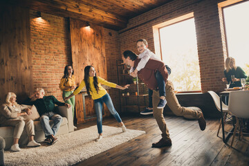 Photo of full big family gathering two little kids play dad piggyback mom laugh daughter plane pose grandparents sit sofa table drink wine talk generation in home evening living room indoors