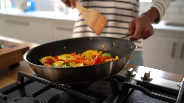 Young woman at home uses wooden spatula to toss colorful bell pepper slices in the frying pan