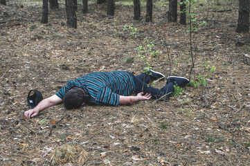 Murder in the woods. The body of a man in a blue t-shirt and trousers lies on the ground among the trees in the forest. Victim of an attack.