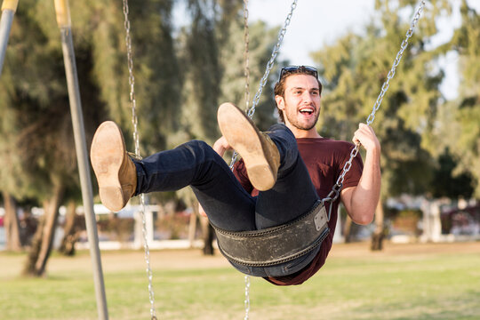 Happy and cheerful young male adult person having fun riding a swing at the park's playground during a sunny summer day.