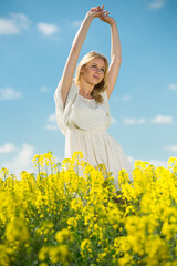 Fototapeta premium Happy girl stretch oneself in rape seed flowers field posing in white dress at sunny day outdoor