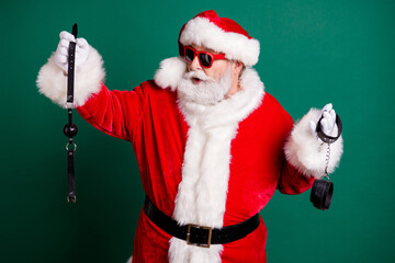 Portrait of his he nice attractive cheery funny Santa grandfather holding in hands choosing whip tools toys equipment passion erotica game theme party isolated over green color background