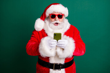 Photo of retired stylish grandpa white beard hold telephone open mouth shocked unexpected sale wear red santa x-mas costume coat gloves sunglass headwear isolated green color background