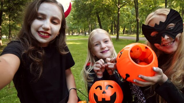 Medium shot of three Caucasian girls wearing costumes of devil, witch and bat are making selfie with pumpkin buckets outdoors