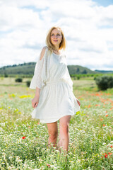 Fototapeta na wymiar Pretty girl wearing white dress walking in field of wild camomile flowers at sunny day