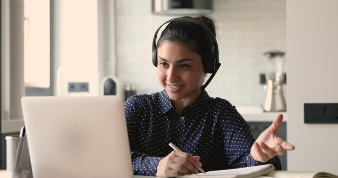 Indian student girl sit at desk at home wear headset talk distantly by video call app and laptop device e-learn language studying with e tutor or classmate do common task remotely, modern tech concept